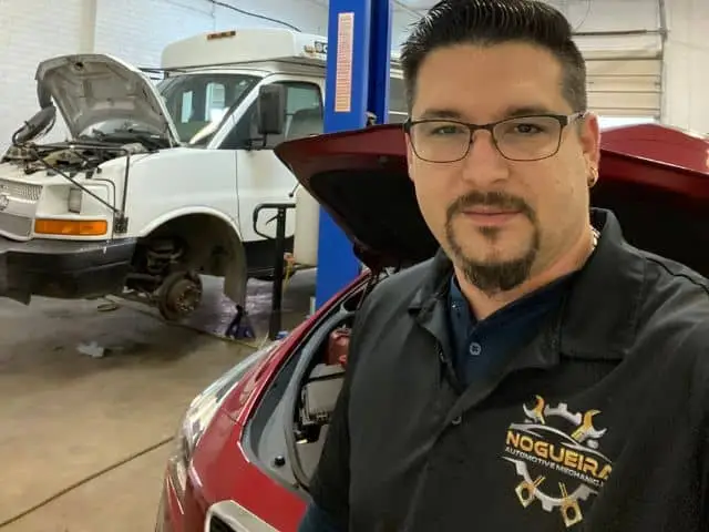 Image of the owner of a repair shop working on two vehicles in the background, holding a tool and smiling